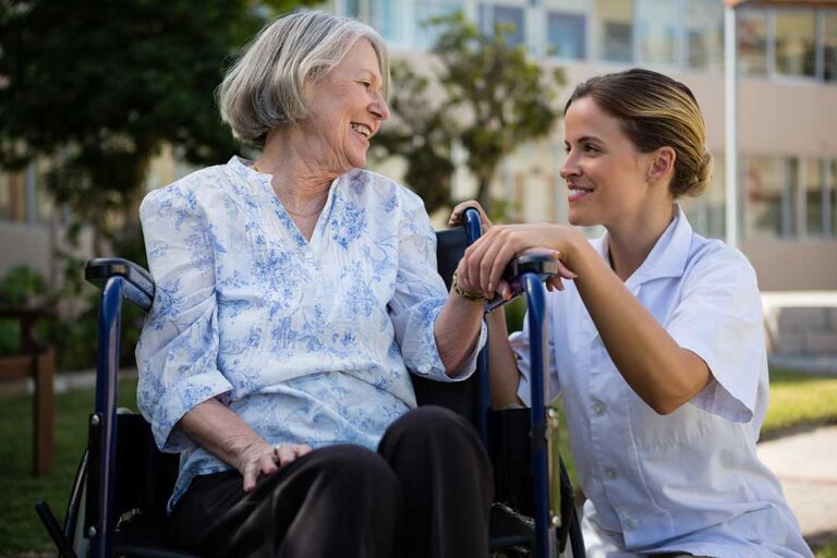 A senior entering a wheelchair-accessible van assisted by a caregiver in front of a residential facility.